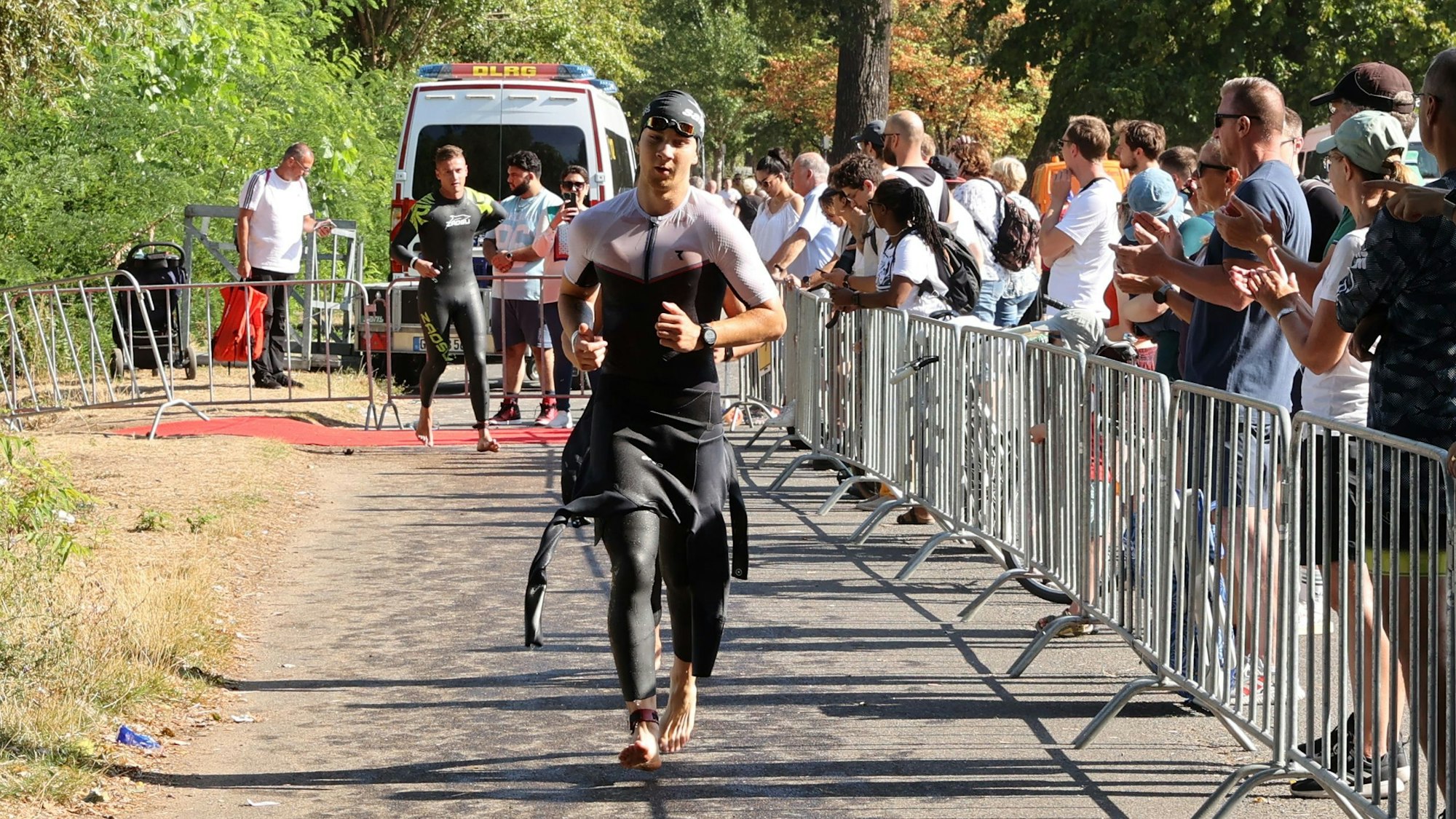 Ein Sportler mit halb ausgezogenem Neoprenanzug läuft über einen Weg. Rechts stehen Zuschauer.