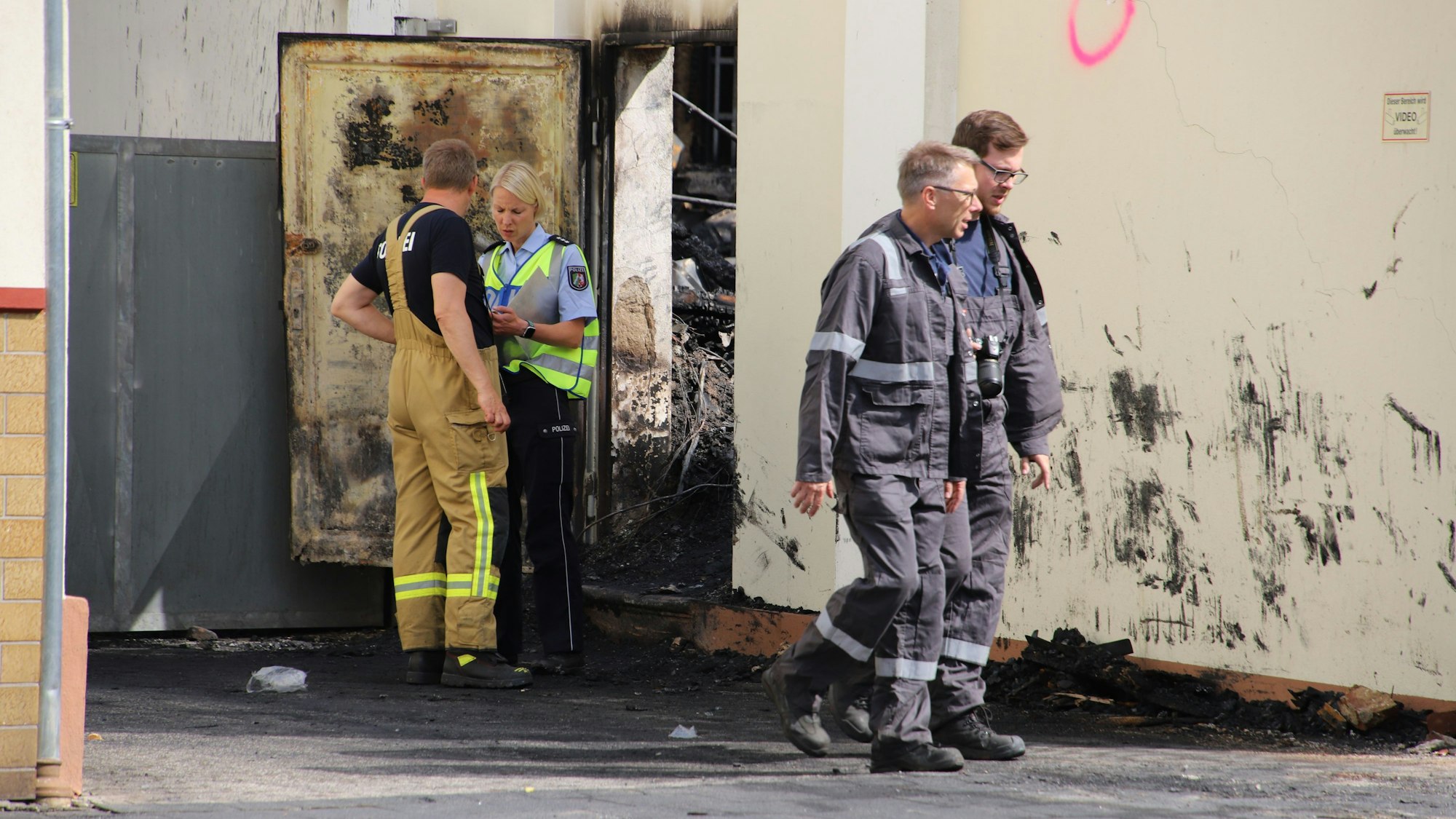 Eine Polizistin und drei Männer in Overalls stehen vor einer verrußten Häuserwand, durch eine Tür sind verkohlte Trümmer zu erkennen.