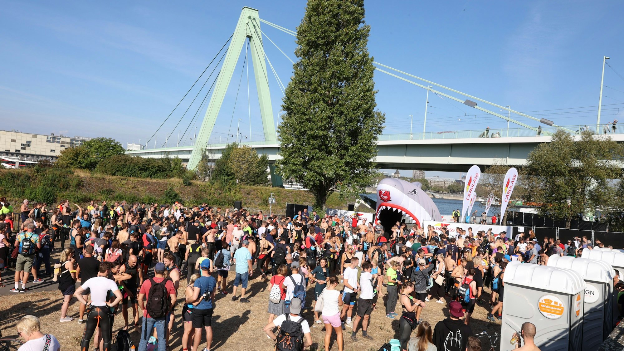 Sportler stehen auf einer Brachfläche unterhalb der Severinsbrücke und warten auf den Schwimmstart des Köln Triathlons.