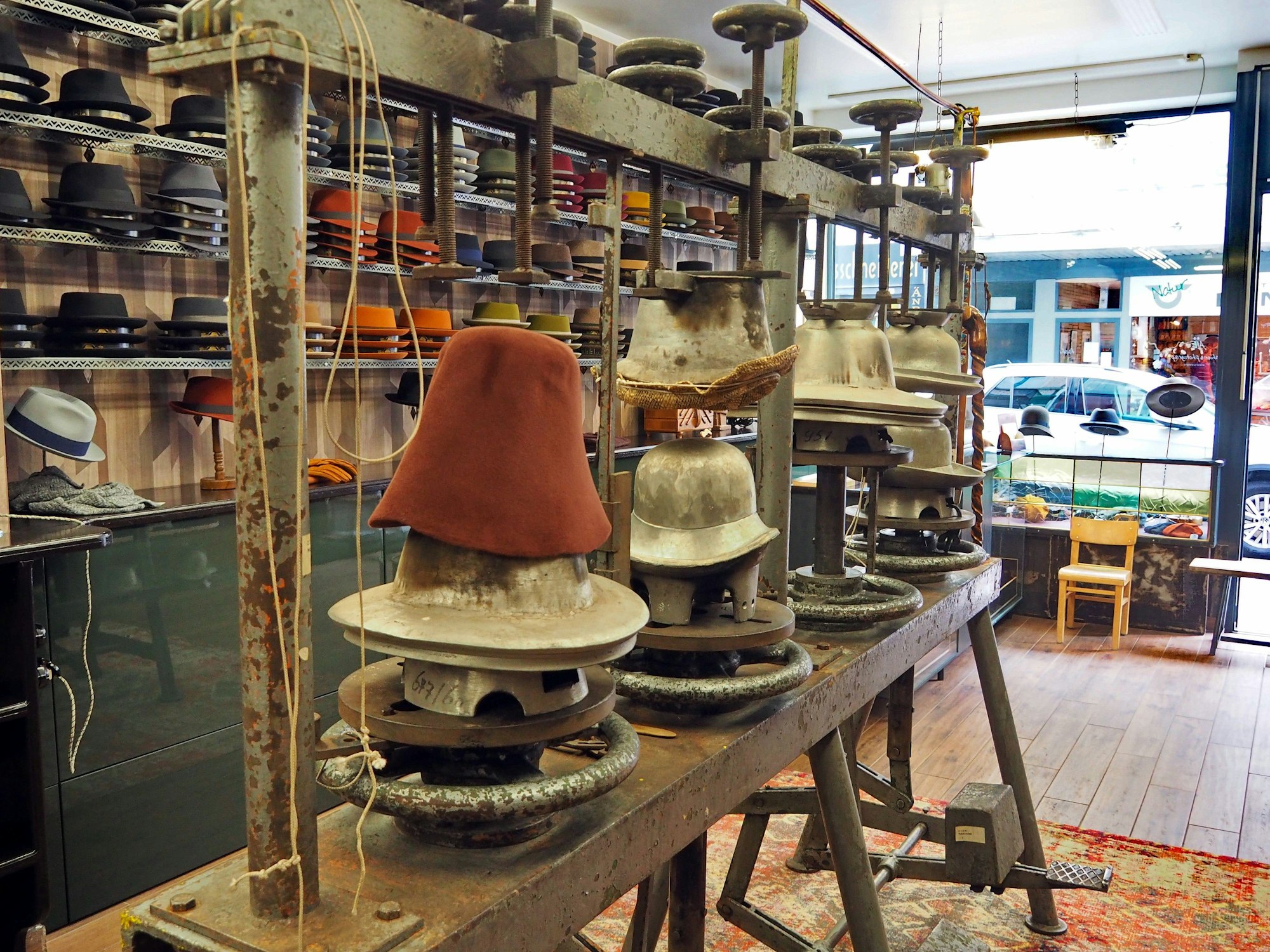 Eine Maschine auf einem Holzgestell, hat verschiedene gusseiserne Formen für Hüte, ein Filzhut ist als Rohling zu sehen.
