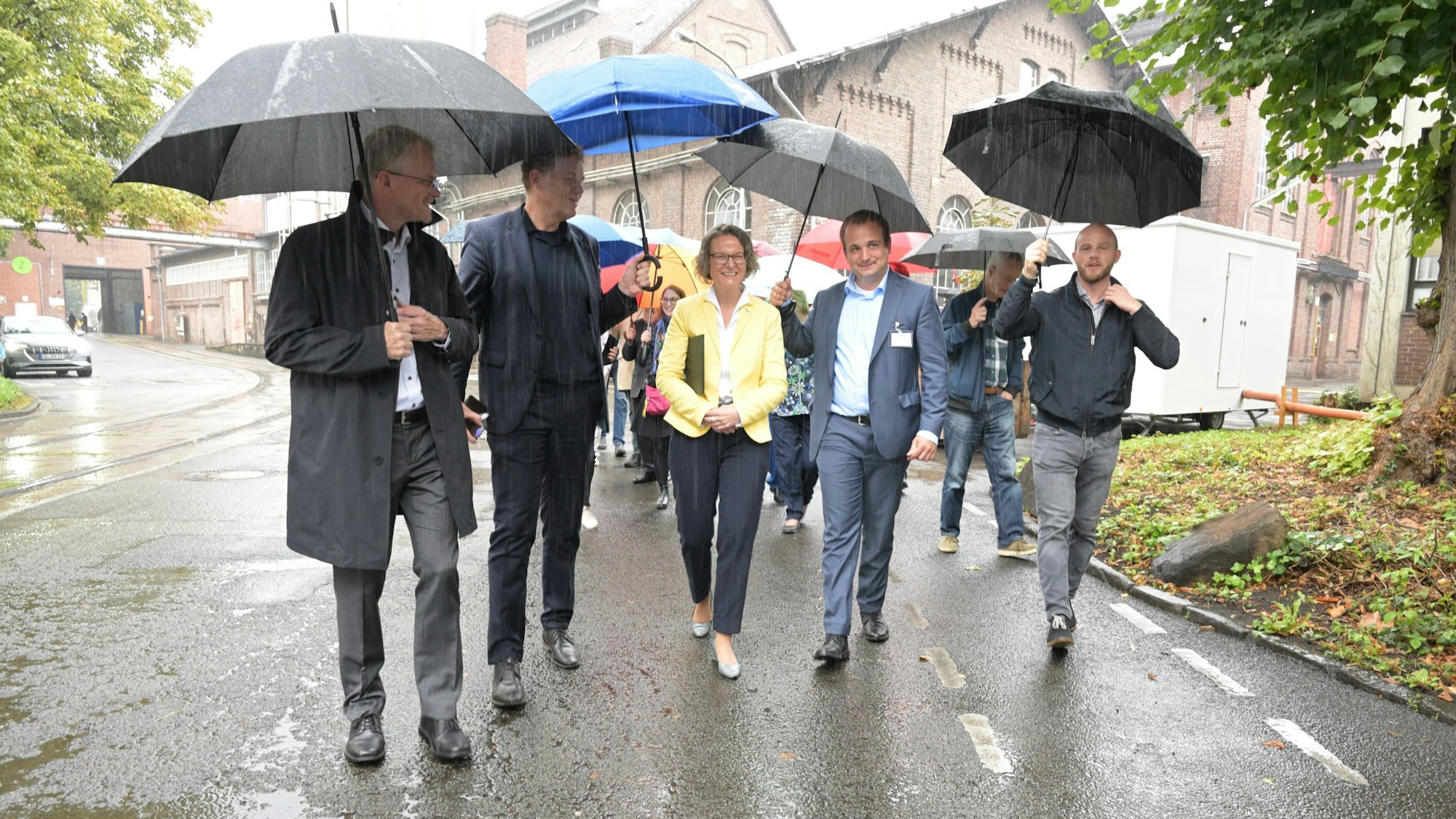 NRW-Bauministerin Ina Scharrenbach (CDU) geht mit lokalen Verantwortlichen unter Regenschirmen übers Zandersgelände.