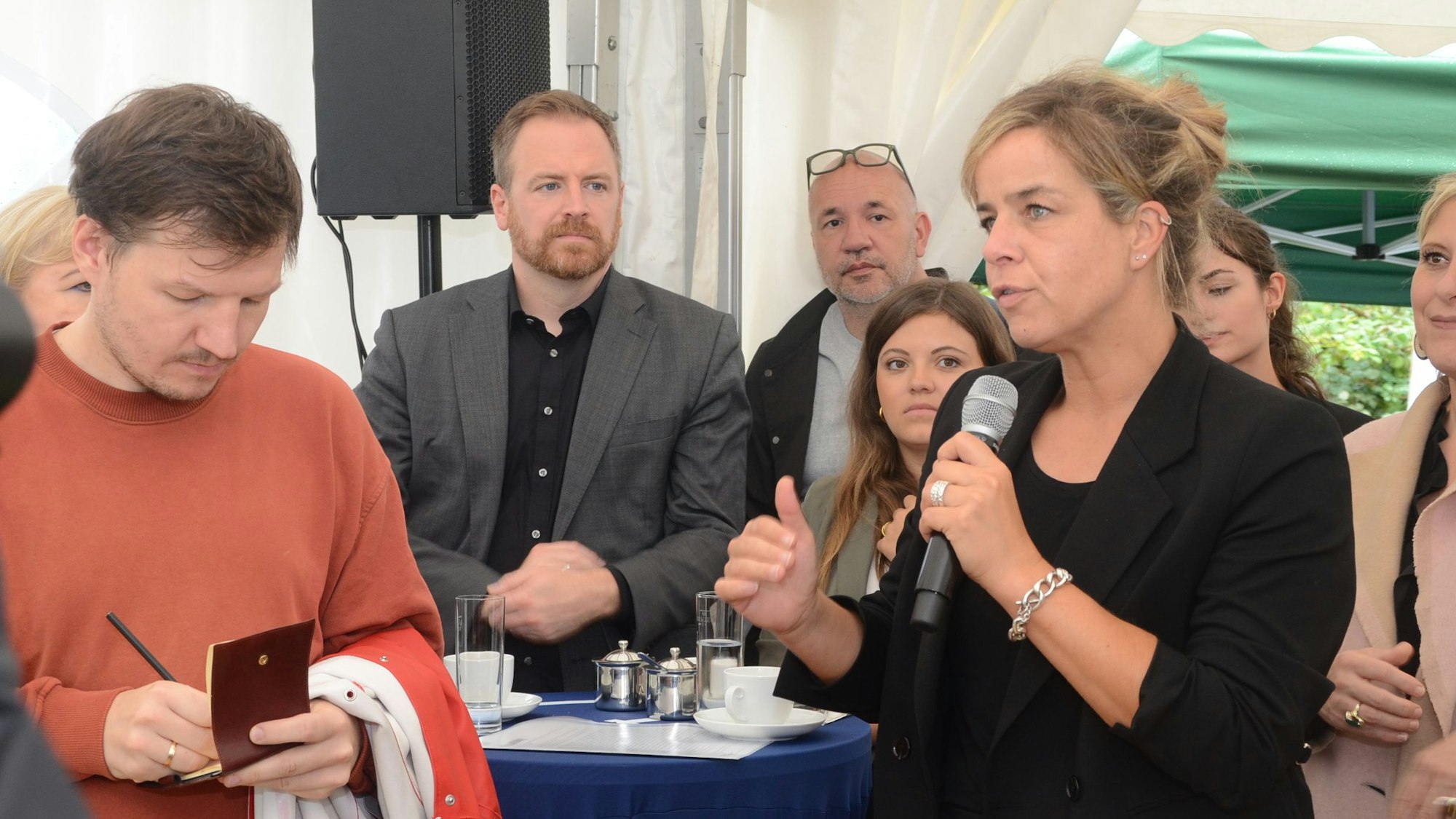 Die Grüne NRW-Wirtschaftsministerin spricht in einem Zelt. Sie hat ein Mikrofon in der Hand. Ein Mann im roten Pullover macht sich Notizen.