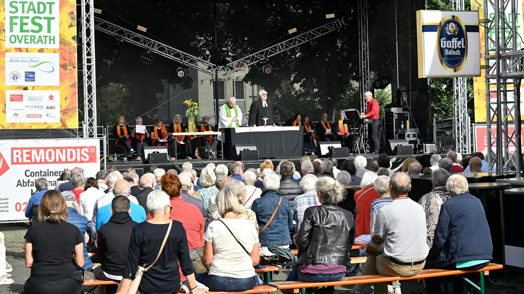 Menschen sitzen auf Bänken und hören den beiden Priestern zu, die auf der Bühne beim Overather Stadtfest einen ökumenischen Gottesdienst zelebrieren.