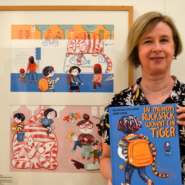 Eine blonde Frau hält das blaue Bilderbuch. Neben ihr hängen zwei Illustrationen aus ihrem Buch, die den katzenähnlichen Tiger mit Kindern zeigen.