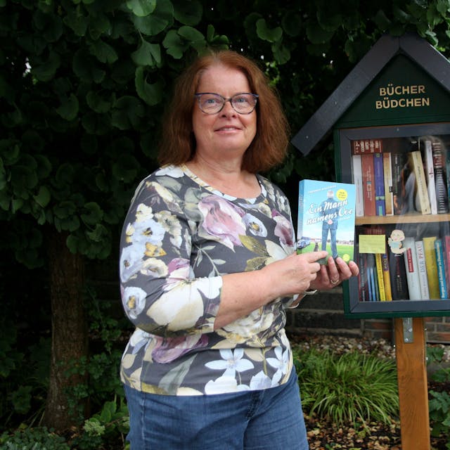 Eine Frau steht mit einem Buch in der Hand vor einem kleinen Bücher-Schrank.