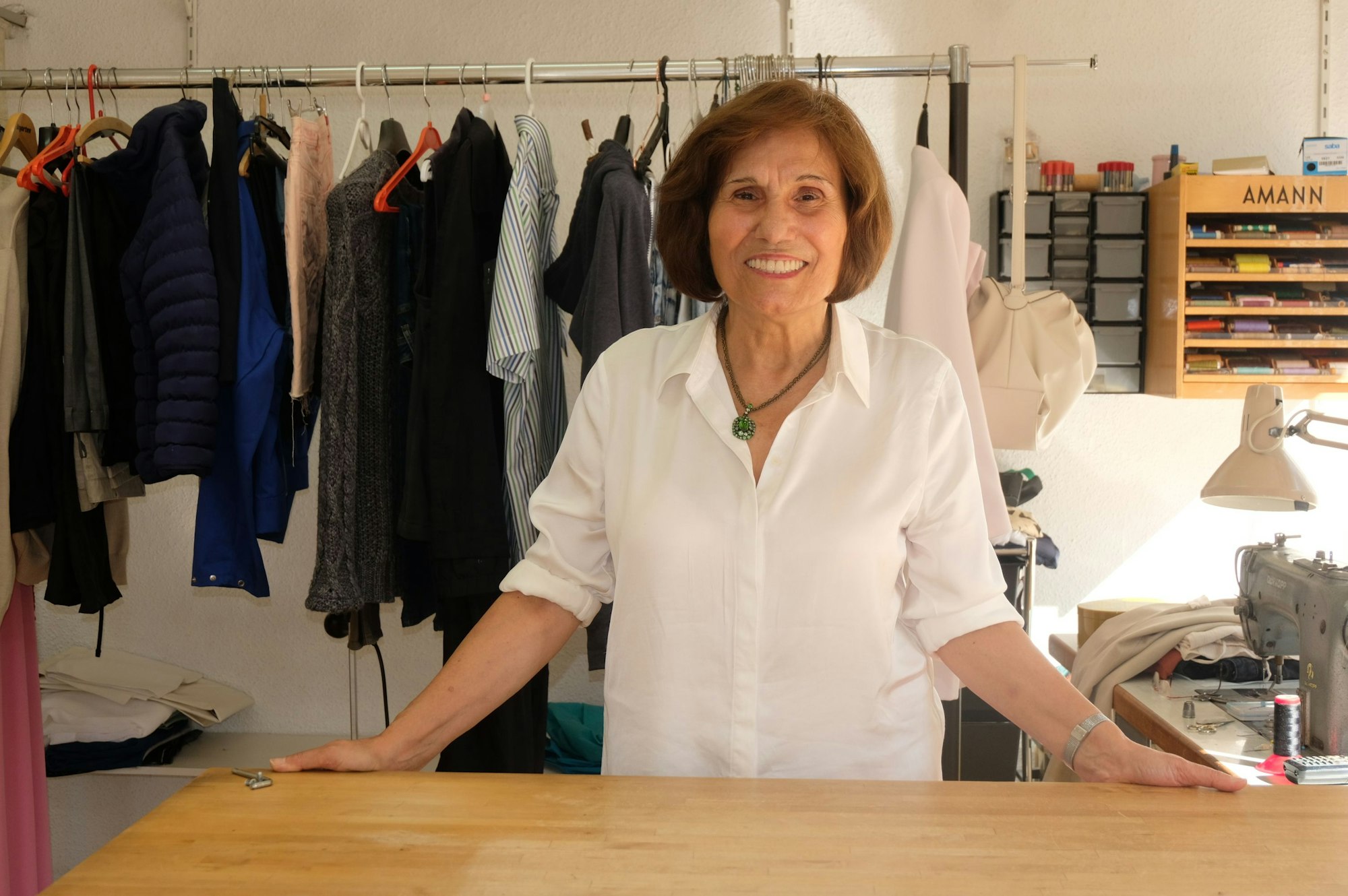 Eine Frau mit brünettem Pagenkopf und einer weißen Bluse steht hinter einer Ladentheke, hinter ihr hängen Kleider, eine Nähmaschine steht auf einem Tisch.