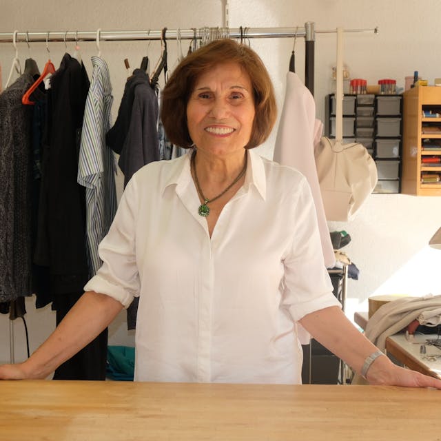 Eine Frau mit brünettem Pagenkopf und einer weißen Bluse steht hinter einer Ladentheke, hinter ihr hängen Kleider, eine Nähmaschine steht auf einem Tisch.