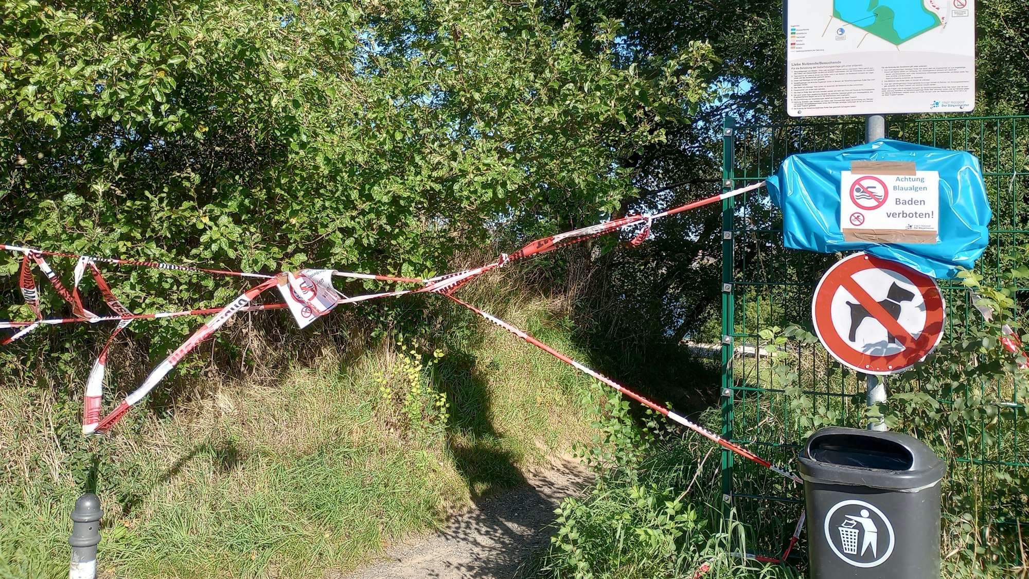 Rot-weißes Flatterband sperrt einen Fußweg ab.An einem Metallpfosten sind eine Karte des Sees, ein Warn- und Verbotsschild und ein Papierkorb befestigt. Auch Hunde sind hier nicht gestattet.