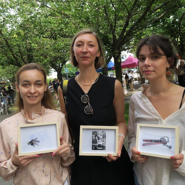 Drei Frauen halten kleinformatige Bilder in der Kamera, hinter ihnen ist ein baumbestandener Platz mit vielen Menschen.