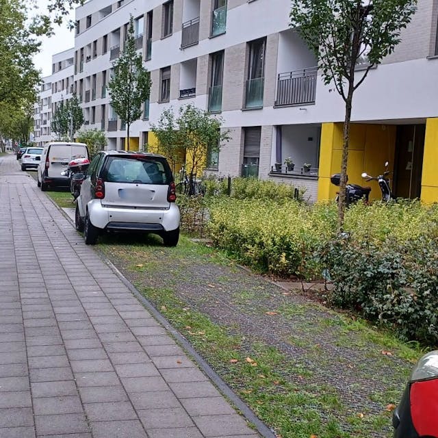Vor einem Wohnblock parken Autos auf einer Schotterfläche neben Rad- und Fußweg.