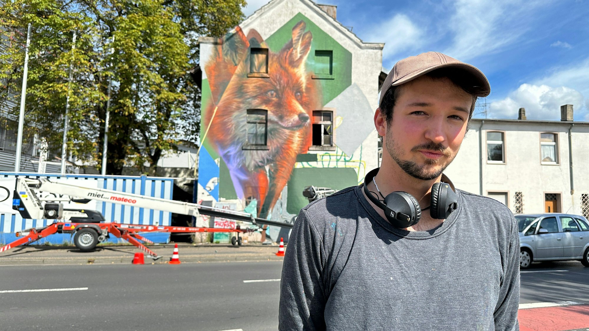 Ein junger Mann mit Käppi und Kopfhörern um den Hals steht an einer Straße. Im Hintergrund ist ein Gebäude zu sehen, auf dem ein großer Fuchs prangt.