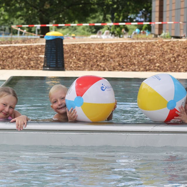Drei Kinder plantschen mit Wasserbällen in einem Schwimmbecken.