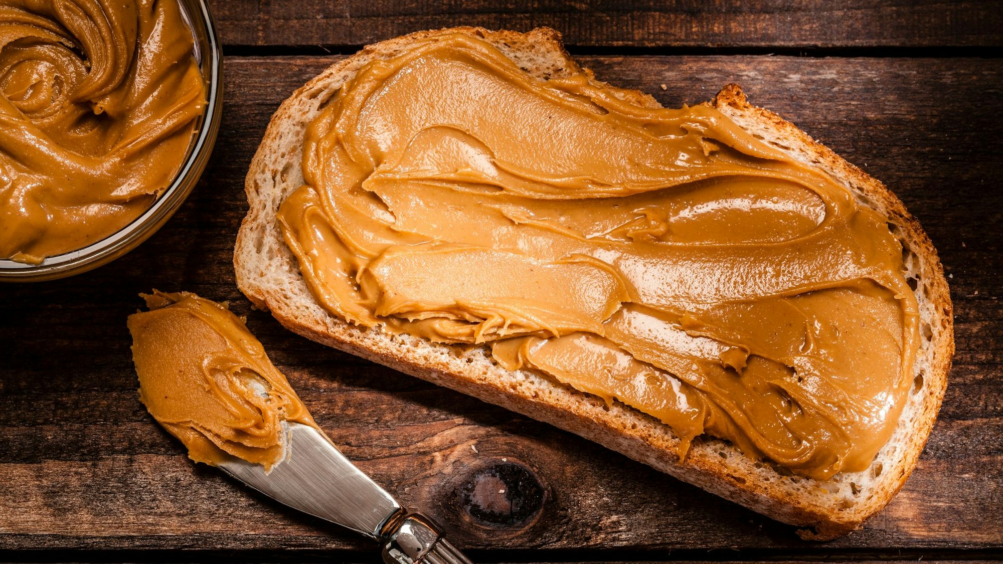 Auf dem Foto sieht man ein geschmiertes Brot mit Erdnussbutter. Daneben liegen das Glas und ein Messer.