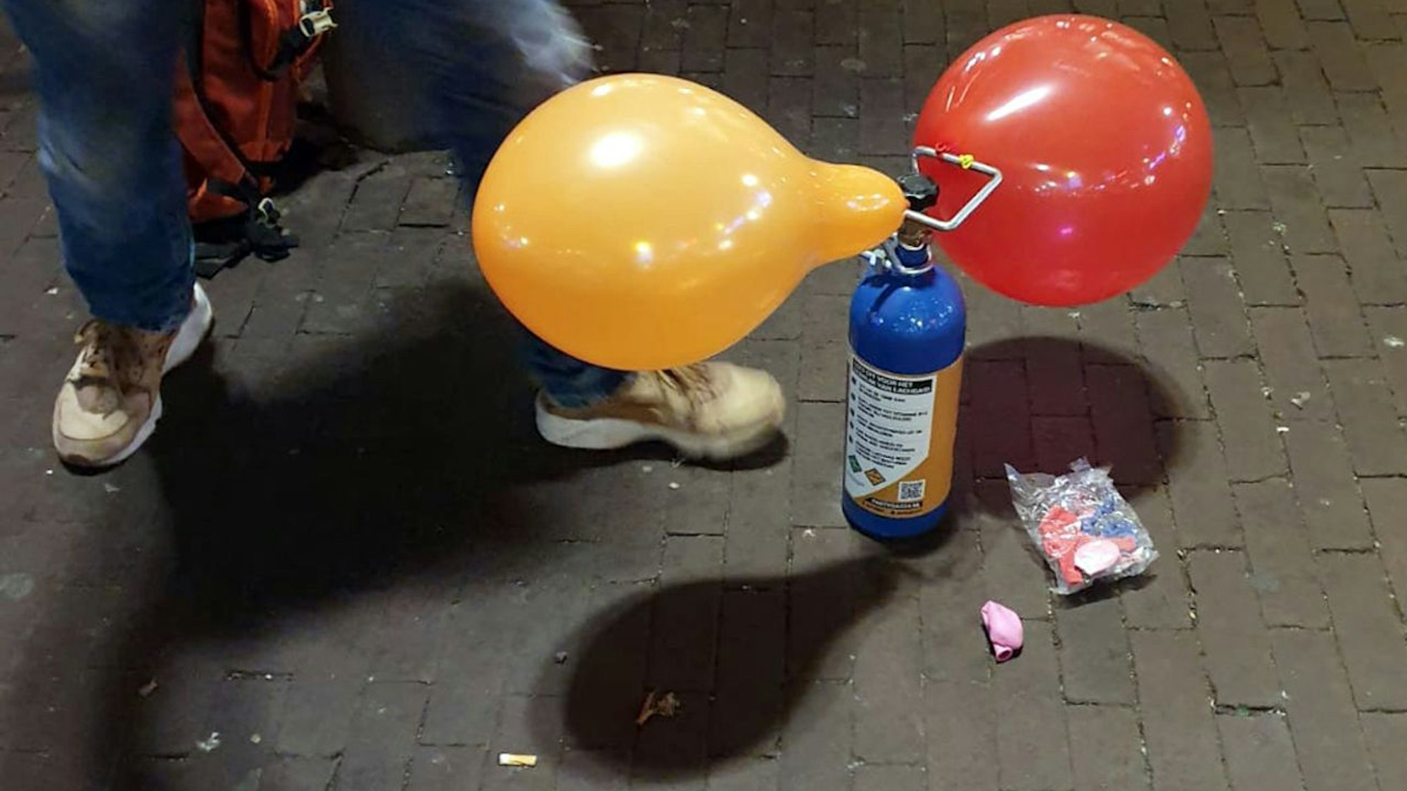 Auf einer Straße steht eine blaue Gaskartusche, an der sich ein gelber und ein roter Luftballon befinden. Die Ballons werden an Ort und Stelle aufgeblasen und die Kunden inhalieren anschließend das Lachgas aus ihnen.