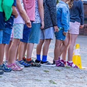 Schüler und Schülerinnen stehen in einer Reihe auf dem Schulhof einer Grundschule. Nur ihre Beine und die unterschiedlichen Schuhe sind zu sehen.