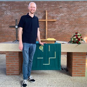Diakon Jan Simons steht vor dem Altar der Evangelischen Kirche in Weilerswist. An der Wand hängt ein hölzernes Kreuz. Auf dem Altar liegen Blumen und die Bibel.