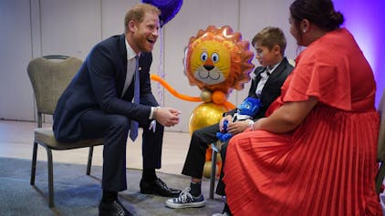 Prinz Harry grinst breit - ihm gegenüber sitzen ein Junge und eine Frau - im Hintergrund befinden sich Luftballons.&nbsp;
