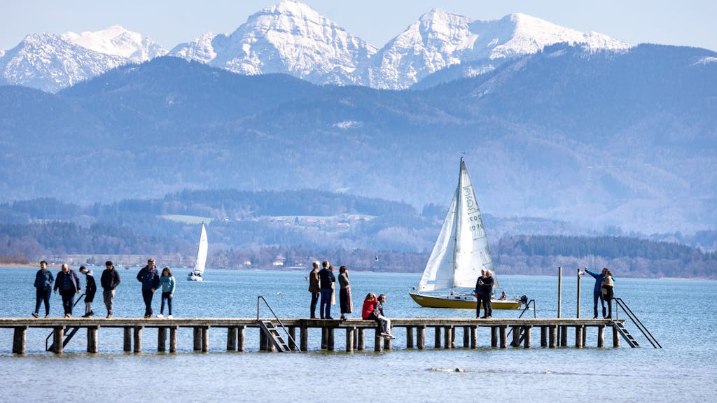 Spaziergänger und Spaziergängerinnen stehen auf einem Steg am Chiemsee, auf dem Segelboote fahren, hier im April 2023 in Bayern.&nbsp;