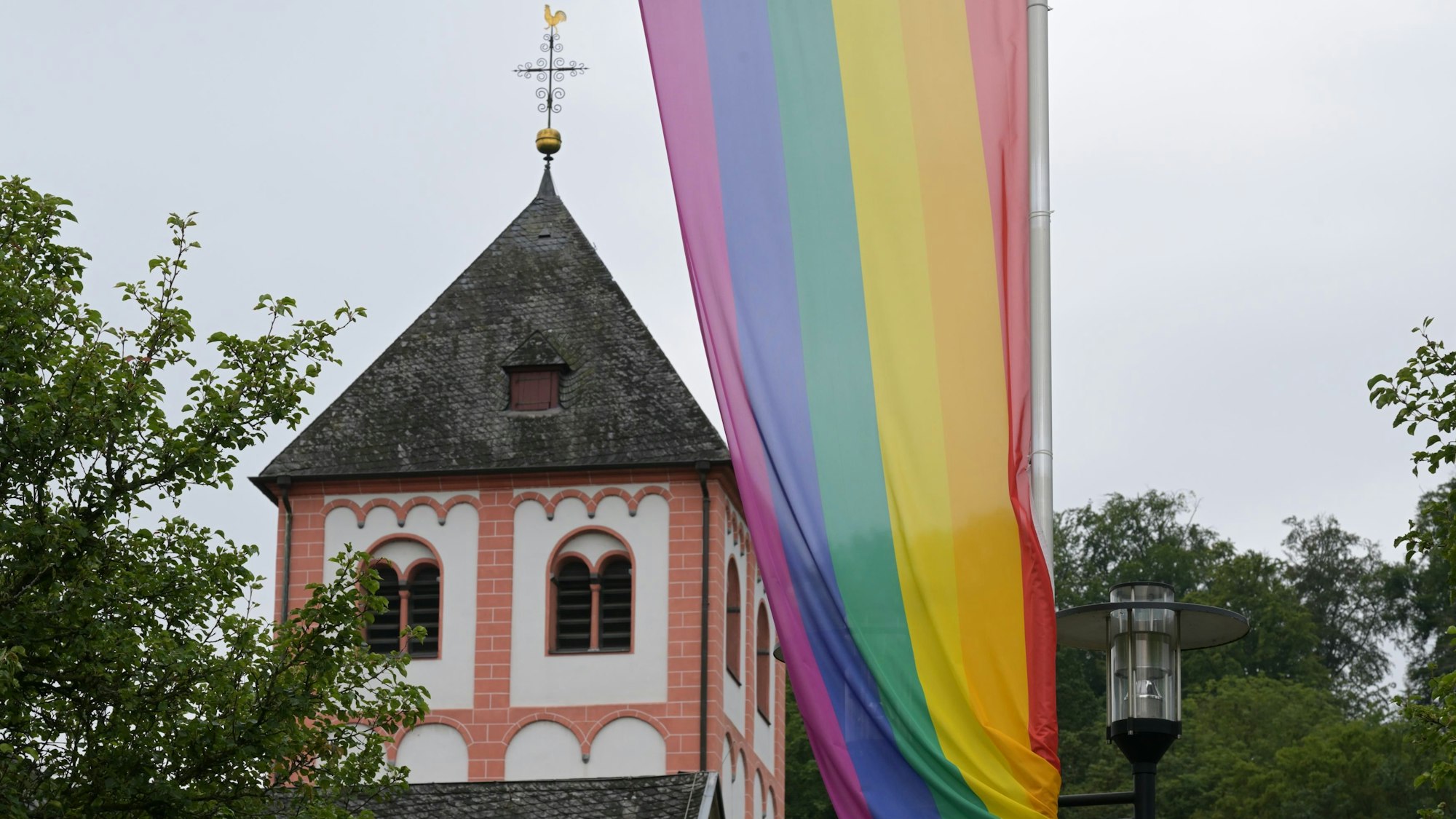 Eine Regenbogenflagge mit sechs Farbstreifen weht im Wind. Im Hintergrund ein Kirchturm.