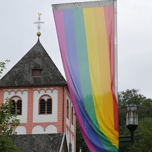 Eine Regenbogenflagge mit sechs Farbstreifen weht im Wind. Im Hintergrund ein Kirchturm.