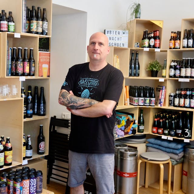 Ein Mann mit Glatzkopf und kleinem Ziegenbart steht in einem Laden, in dem die Regale vollgestellt sind mit Bierflaschen.