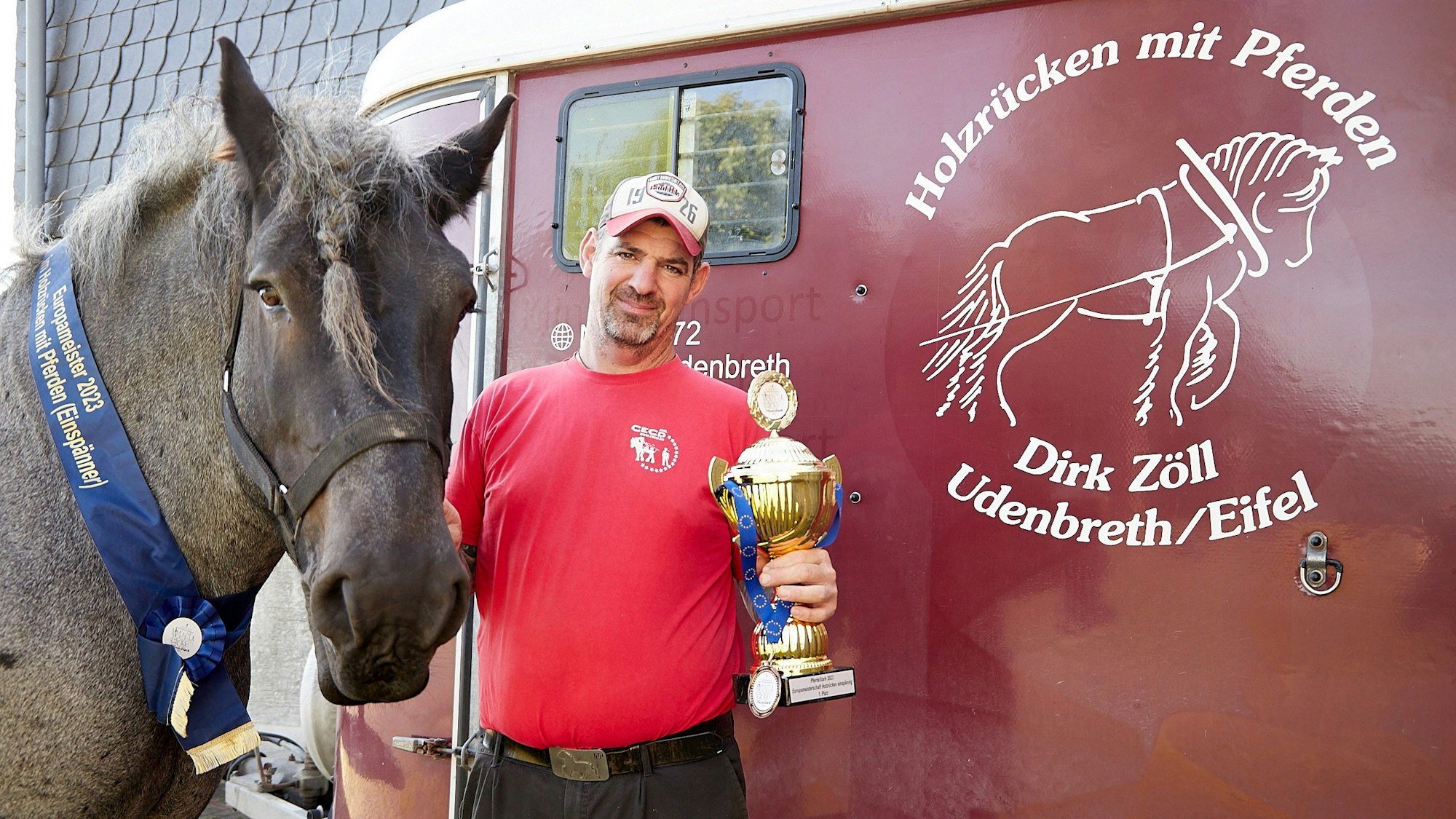 Dirk Zöll vor seinem Pferdeanhänger. In der einen Hand hält er den Europameisterpokal, mit der anderen hält er sein Pferd Wotan fest, dass eine Medaille um den Hals trägt.