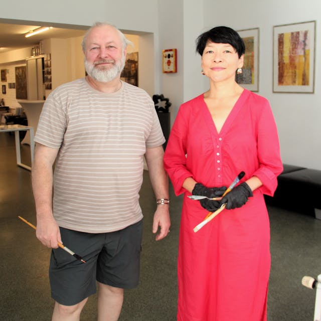 Ein Mann mit grauem Haar und Bart stehe neben einer Frau im roten Kleid mit kurzem schwarzem Haar. Beide haben Pinsel in der Hand.