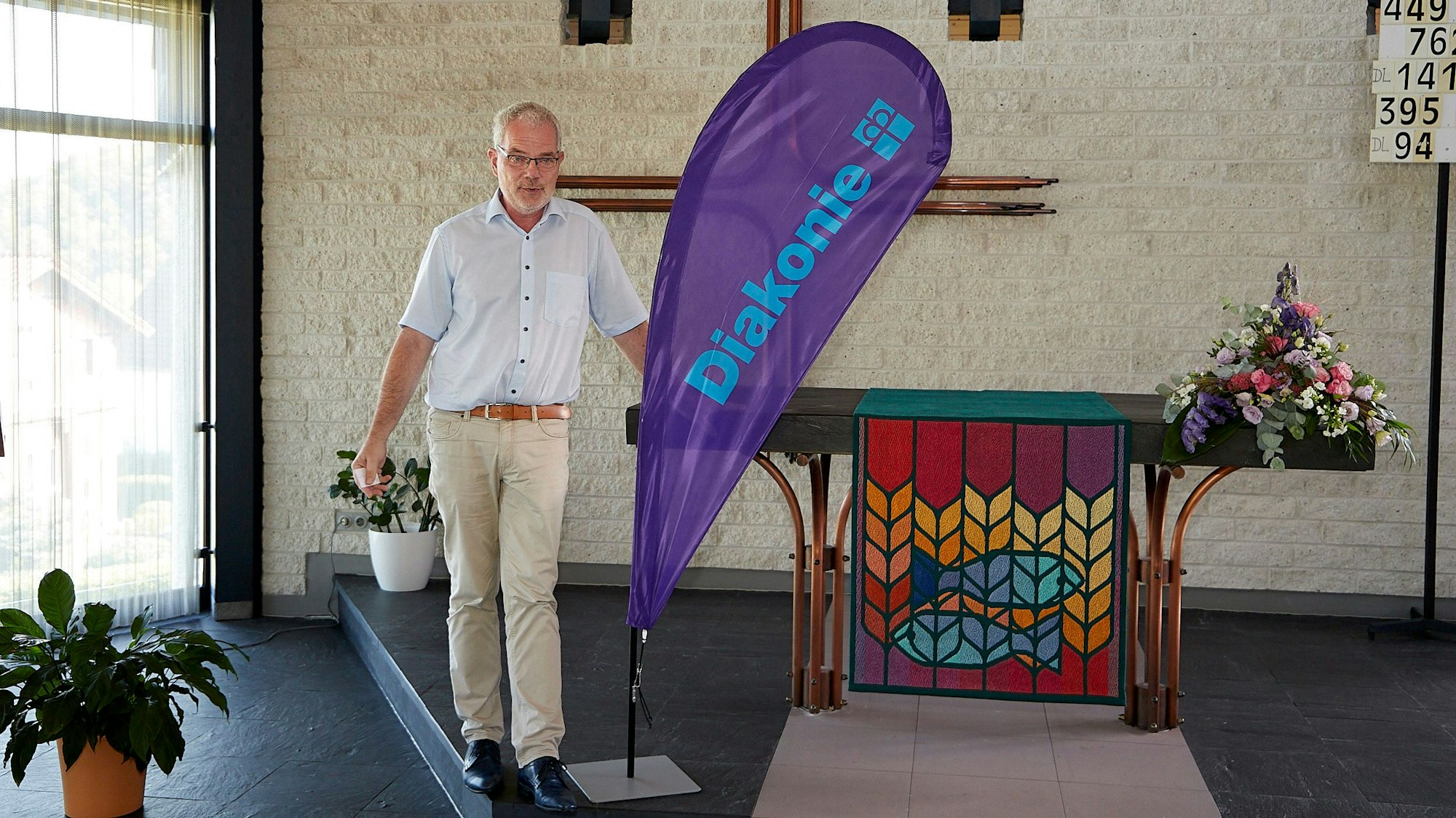 Malte Duisberg steht am Alter der entwidmeten Kirche und begrüßt an einer lilafarbenen Diakonie-Fahne die Anwesenden als neuer Hausherr.