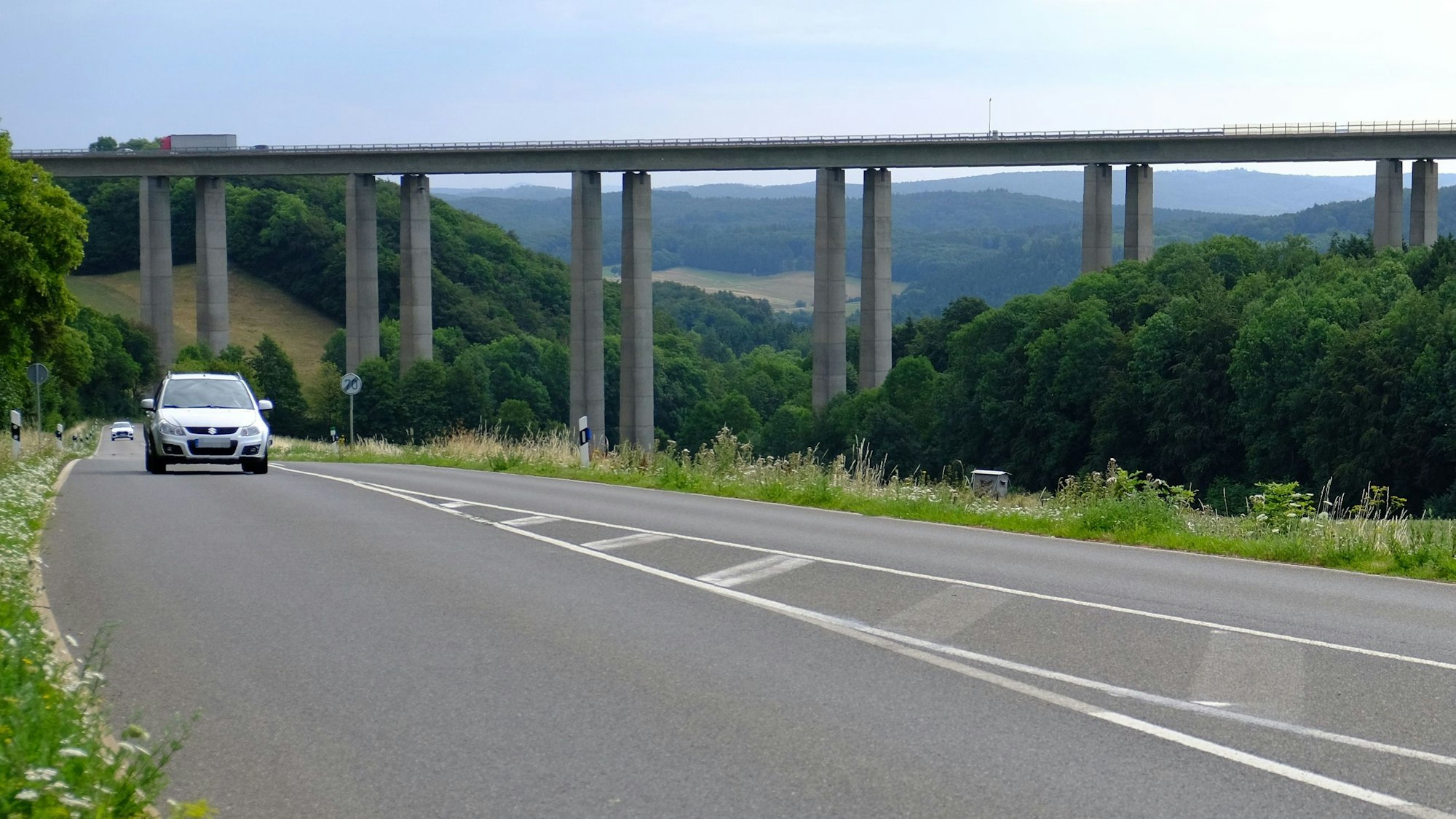 Das Bild zeigt die Talbrücke Zingsheimer Wald der Autobahn 1. Auf einer Landstraße, die unter der Brücke verläuft, sind zwei Autos unterwegs.