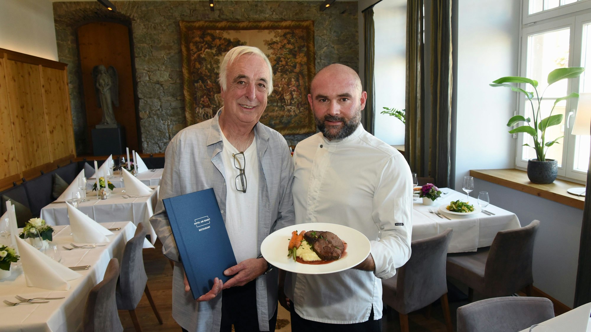 Klaus Ortlieb und Armend Mustafi stehen im Gastraum des „Hotels am Markt“ in Wipperfürth und zeigen ein Gericht von der neuen Speisekarte.