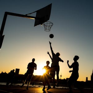 Männer spielen im Licht der untergehenden Abendsonne Basketball am Rheinufer.