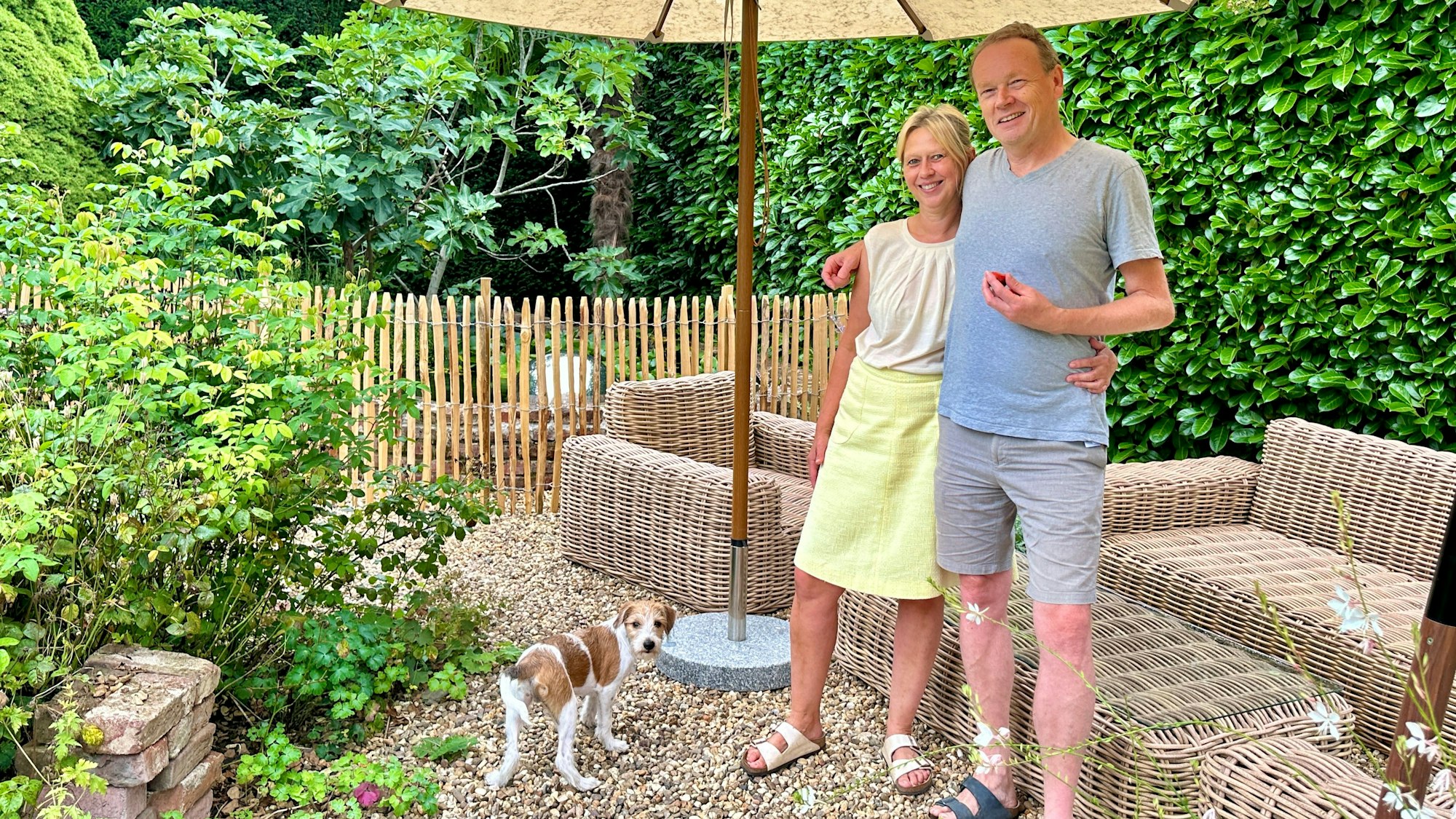 Ein Paar steht in einem Garten vor einer Draußen-Sitzgarnitur aus Korbgeflecht. Sie stehen unter einem Sonnenschirm, ein weißer Hund mit braunen Flecken schaut in die Kamera.