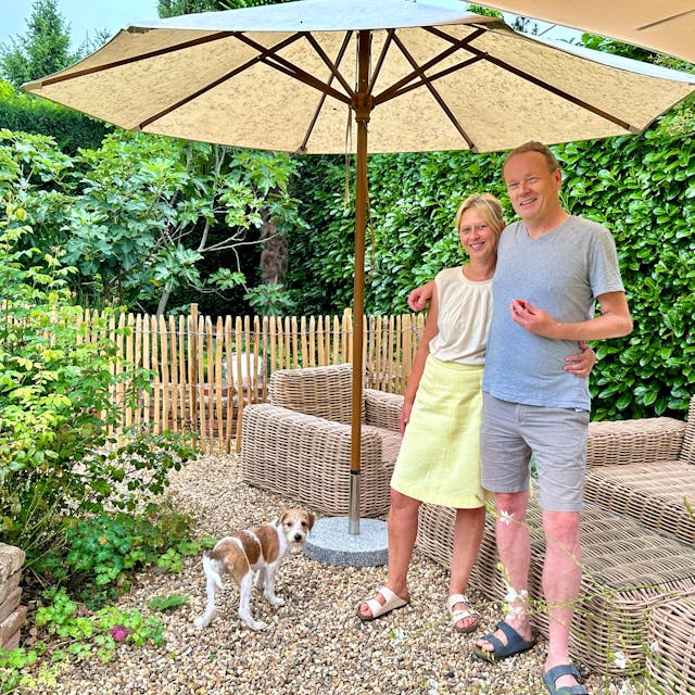 Ein Paar steht in einem Garten vor einer Draußen-Sitzgarnitur aus Korbgeflecht. Sie stehen unter einem Sonnenschirm, ein weißer Hund mit braunen Flecken schaut in die Kamera.