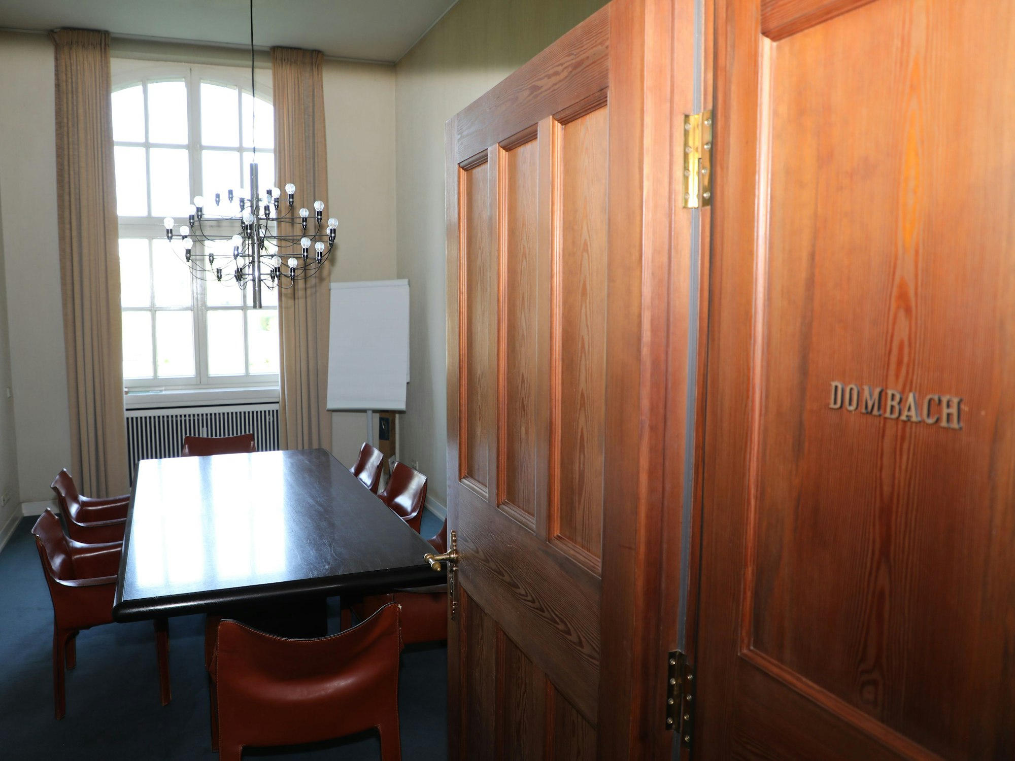 Durch eine offene Tür schaut man in einen Raum mit Besprechungstisch. An der Tür steht „Dombach“ in Messinglettern.