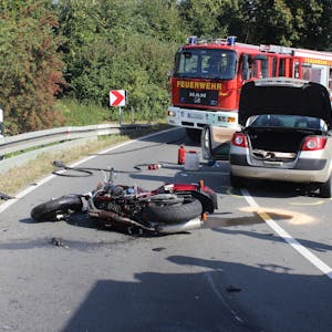 Ein Motorrad liegt auf der Fahrbahn, dahinter der Unfall-Pkw und zwei Einsatzfahrzeuge der Feuerwehr.