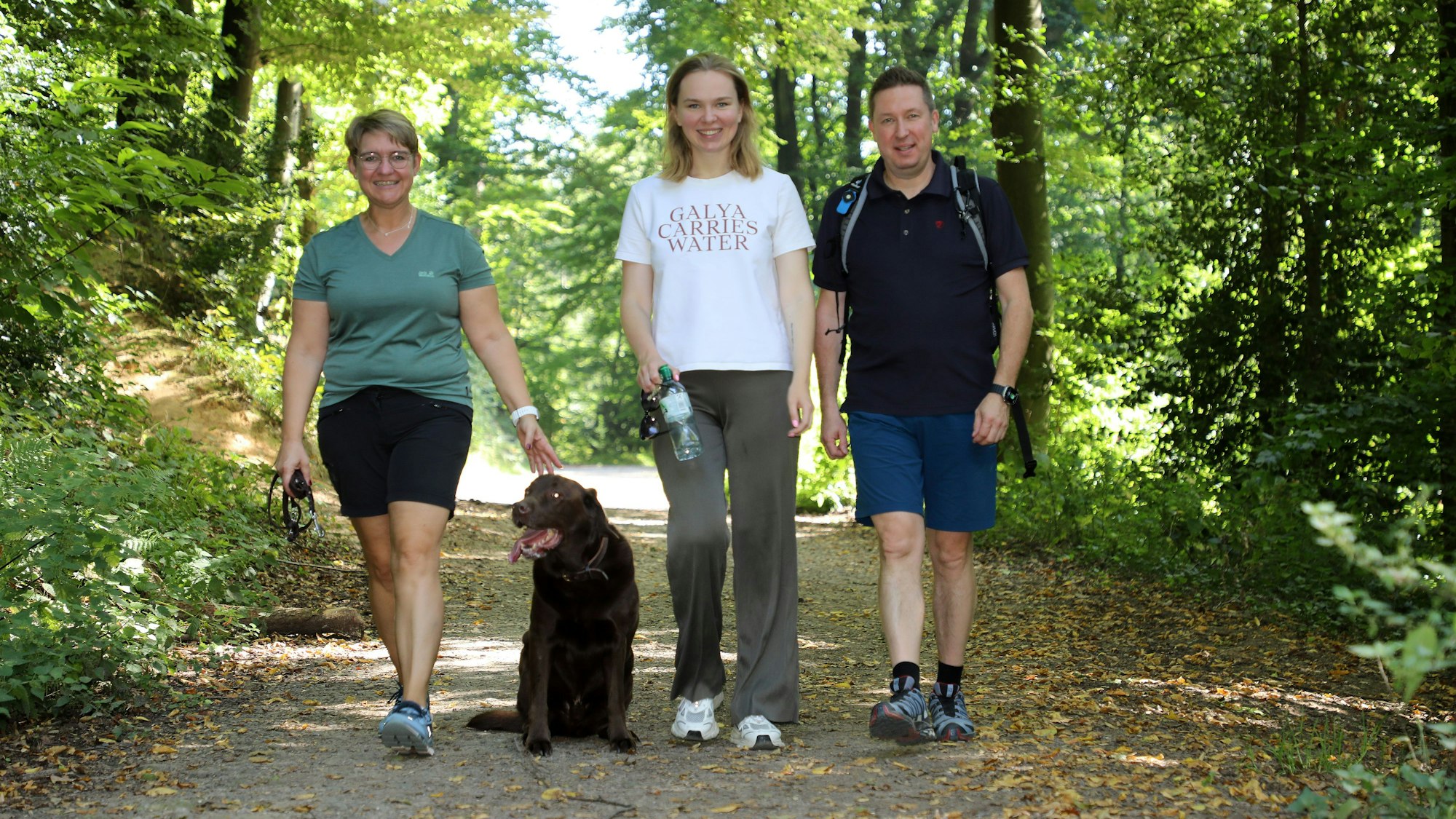 Drei Menschen mit einem Hund wandern auf einem breiten Weg durch den Wald bei Bergisch Gladbach.