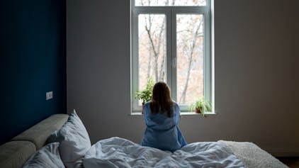 Eine Frau sitzt mit dem Rücken zur Kamera auf einem Bett und blickt aus dem Fenster.