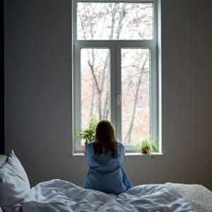 Eine Frau sitzt mit dem Rücken zur Kamera auf einem Bett und blickt aus dem Fenster.