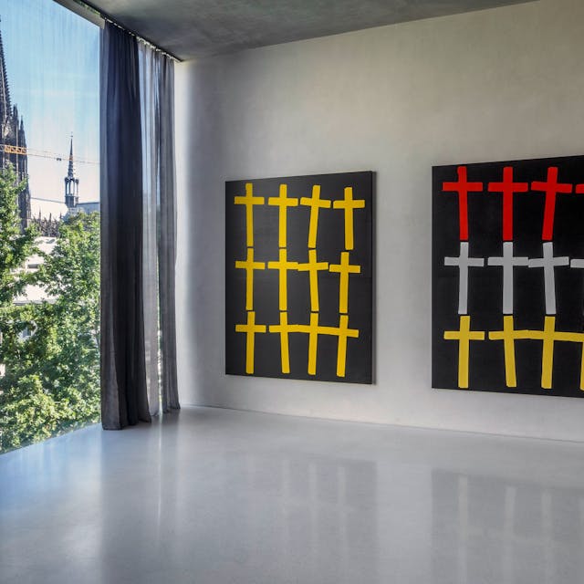 Zwei Bilder mit Kreuzen hängen neben einem Fenster, durch das der Kölner Dom zu sehen ist.