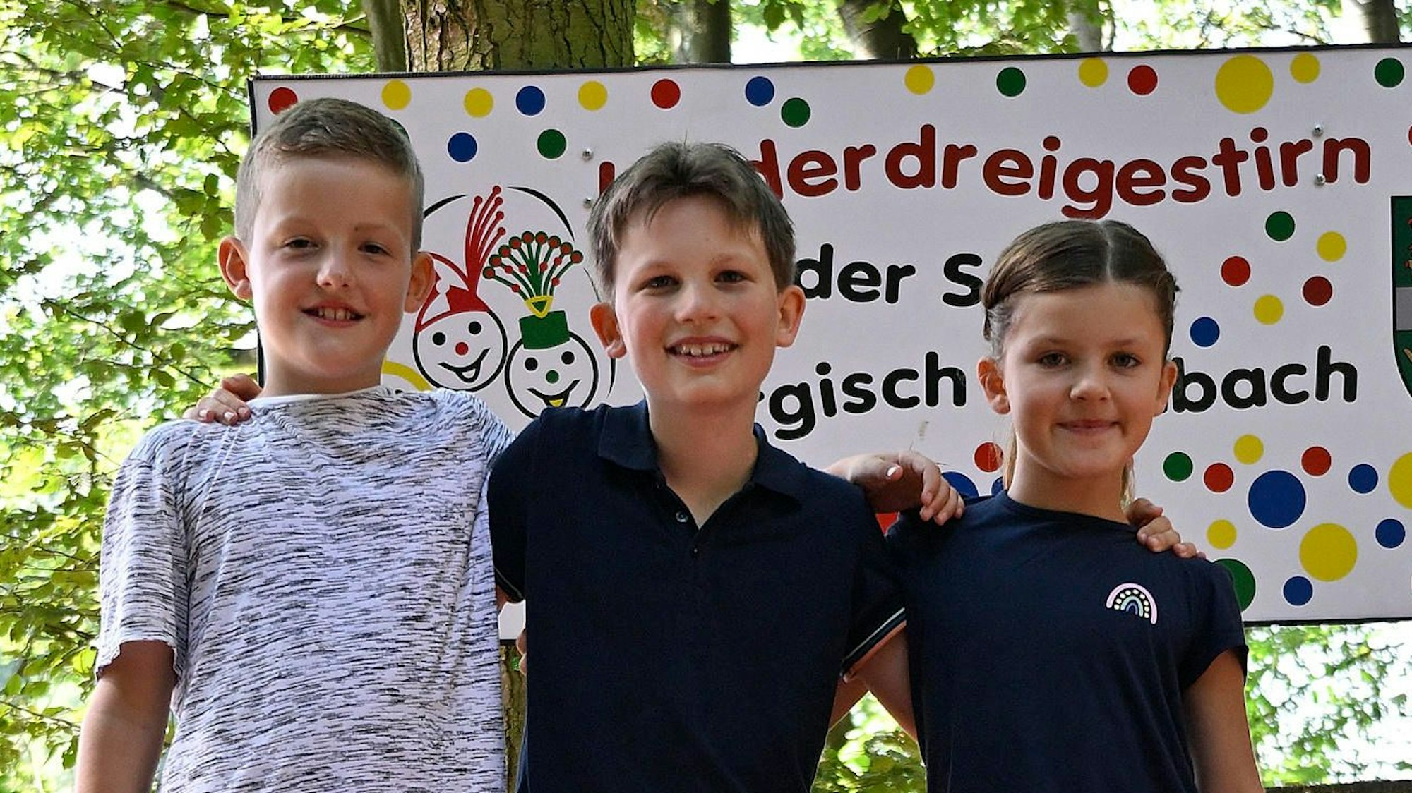 Das angehende Kinderdreigestirn der Kreisstadt Bergisch Gladbach steht im Wald vor einem Schild mit der Aufschrift „Kinderdreigestirn der Stadt Bergisch Gladbach“.