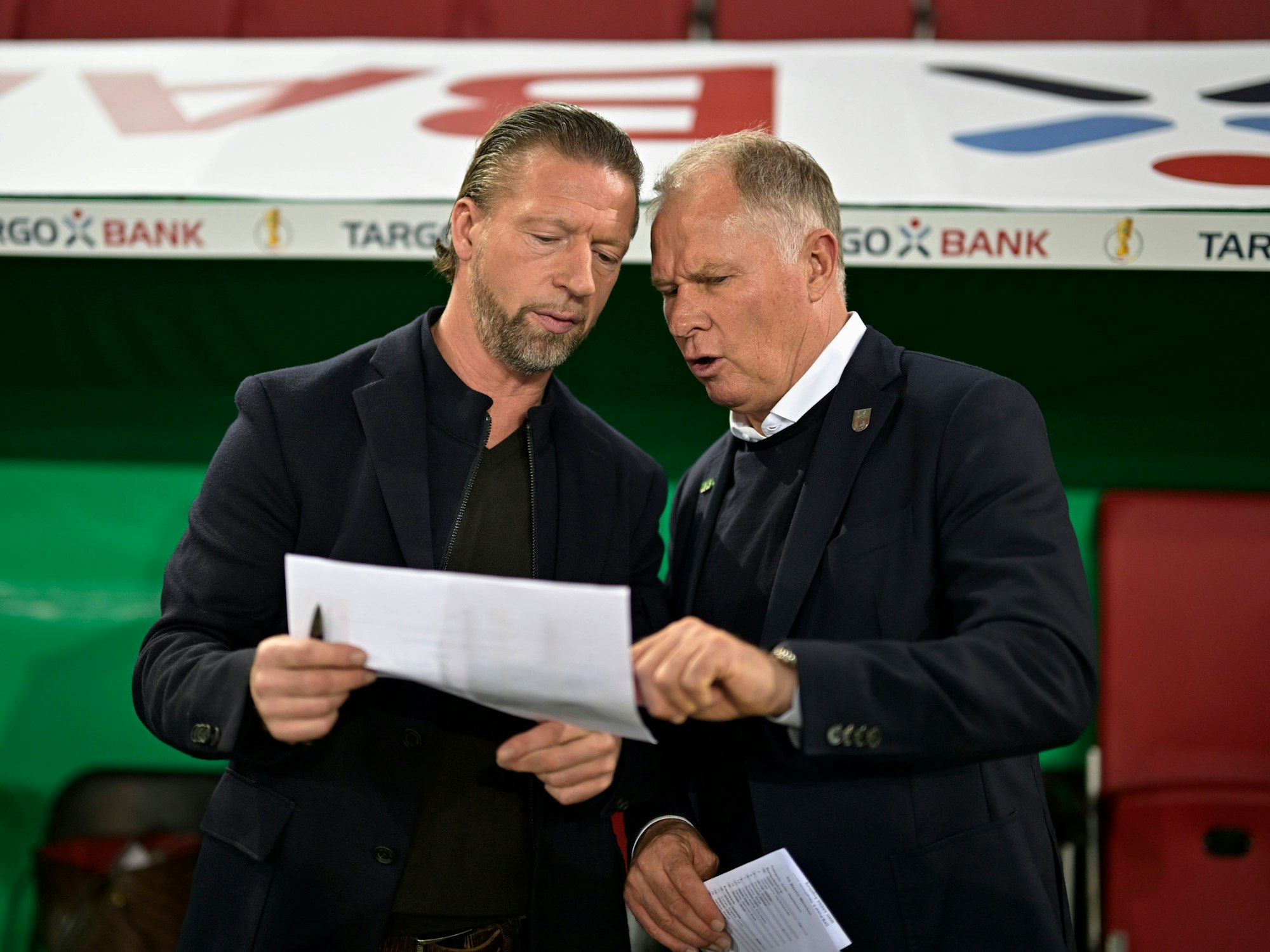 Steffen Freund unterhält sich mit Stefan Reuter. Freund hält einen Zettel in den Händen, auf den beide schauen.