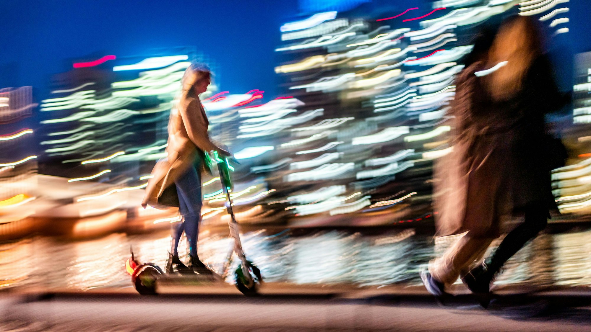 Vor den erleuchteten Hochhäusern sind am Abend eine Frau auf einem E-Scooter (l) und zwei Spaziergängerinnen (r) unterwegs. Das Bild hat einen starken Wischeffekt.