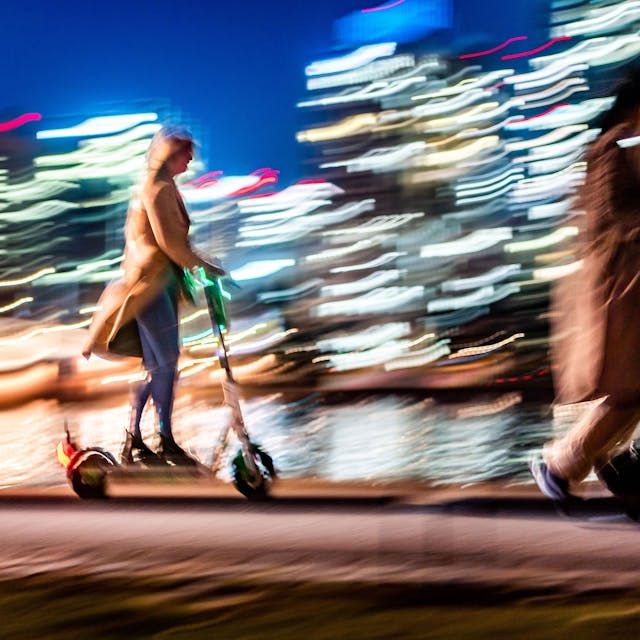 Vor den erleuchteten Hochhäusern sind am Abend eine Frau auf einem E-Scooter (l) und zwei Spaziergängerinnen (r) unterwegs. Das Bild hat einen starken Wischeffekt.&nbsp;