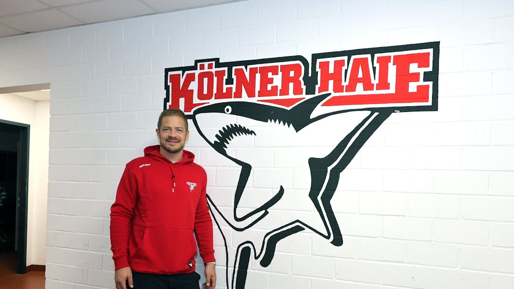 Moritz Müller (Haie) steht vor dem Logo des KEC in der Arena-Kabine.