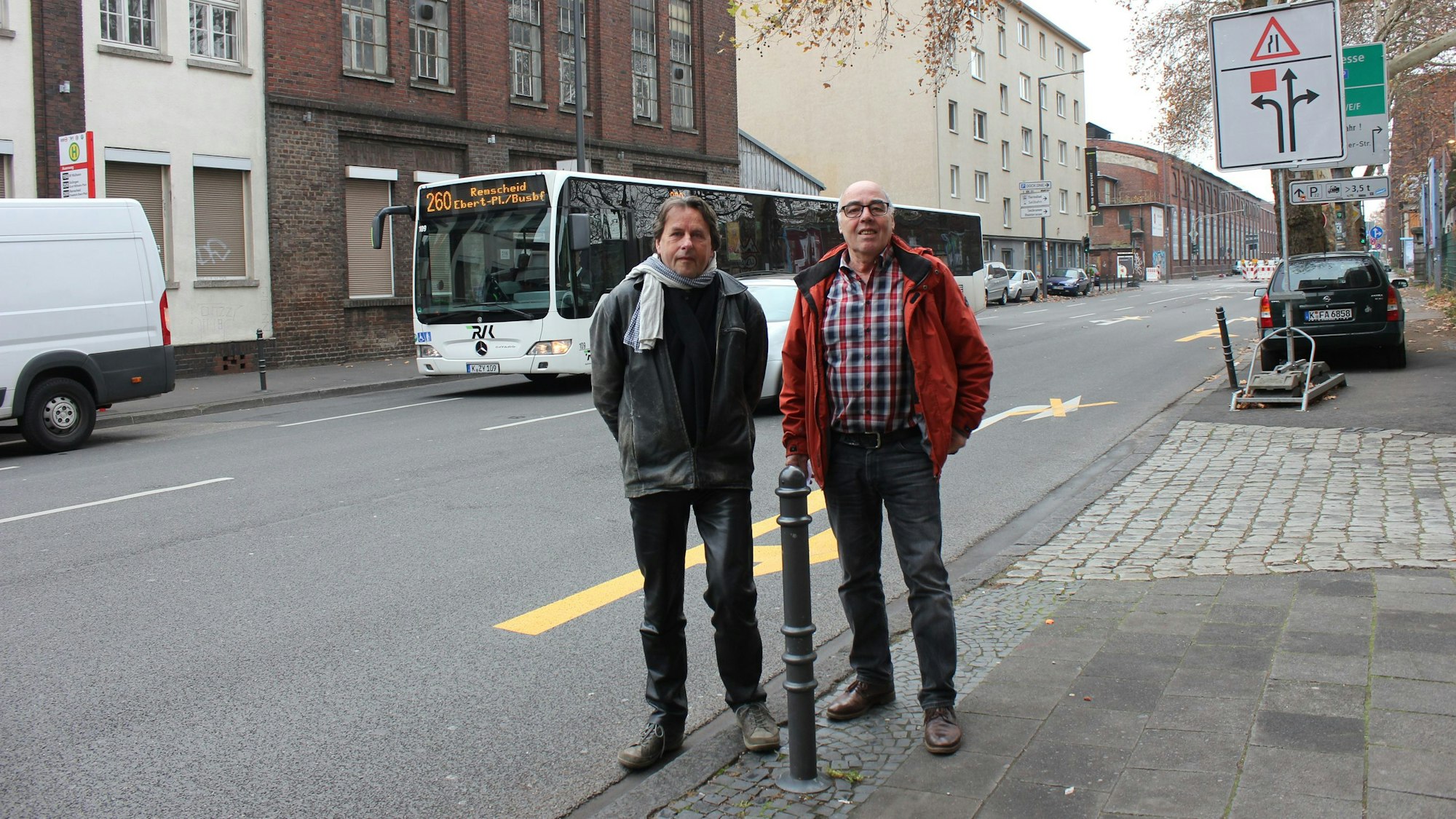 Zwei Männer stehen nebeneinander an einer Straße, hinter ihnen ist ein Bus zu sehen.