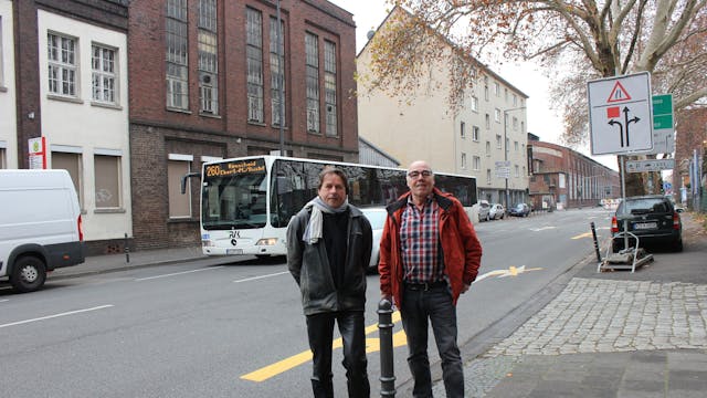 Zwei Männer stehen nebeneinander an einer Straße, hinter ihnen ist ein Bus zu sehen.&nbsp;