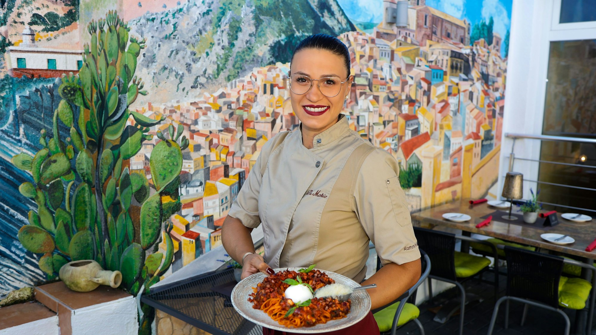 Köchen Ramona Vella-Bianchettino zeigt im Vordergrund ein Pasta-Gericht, im Hintergrund ein Wandbild der sizilianischen Stadt Siculiana.