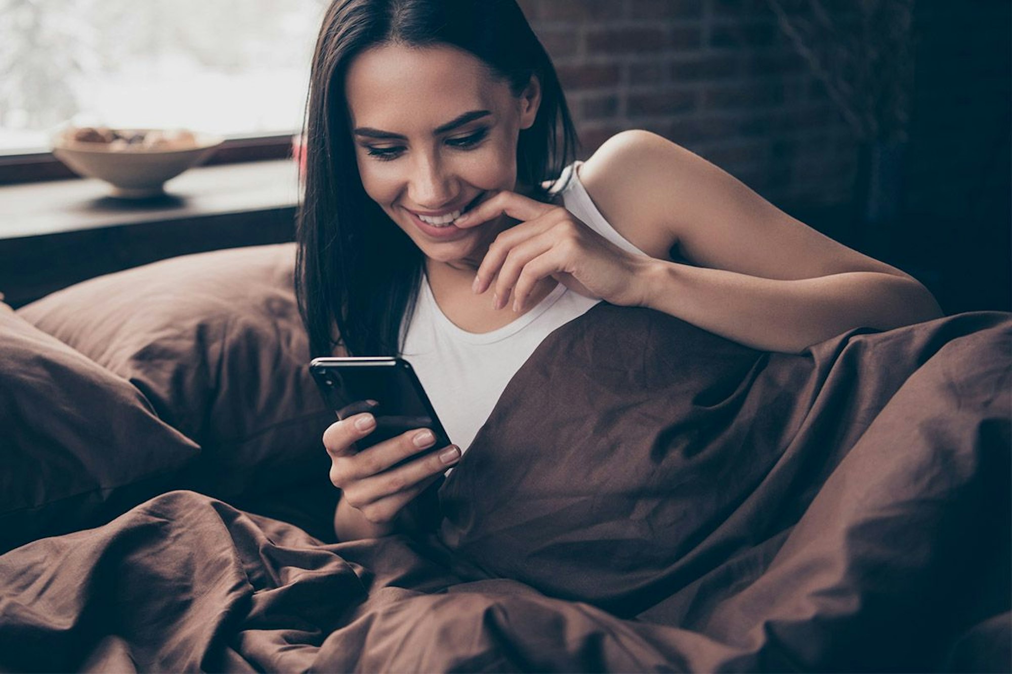 Frau im Bett schaut freudig gespannt auf ihr Handy.