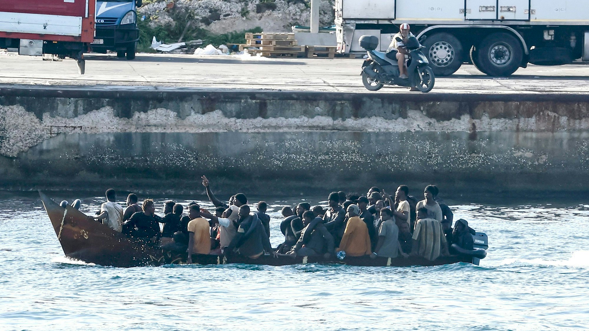 Ankunft von Migranten auf Lampedusa: Ein überbesetztes kleines Fischerboot mit Außenbordmotor läuft in einen Hafen ein.