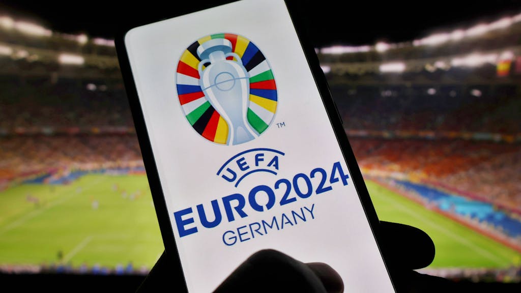 Das Display eines Handys zeigt das Logo der EM 2024 in Deutschland. Im Hintergrund ist der Innenraum eines Stadions zu